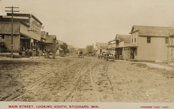 Text on front reads: "Main Street, Looking South, Stoddard, Wis." An unpaved main street with businesses on both sides. On the left is a store with a sign that reads: "Hardware. 5 & 10¢ Store." It has rolled up fencing with various implements and merchandise on the porch. A horse and wagon are hitched in front. The name "Blashek" is hand written on the storefront. More horse-drawn wagons are in the street. On the right men gather on a porch and a dog rests on another porch. Trees and hills are in the distance.