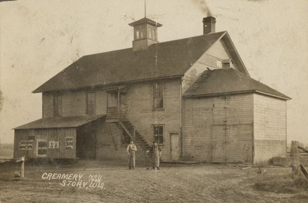 Text on front reads: "Creamery, Story, Wis." Two men pose in front of a two-story, clapboard creamery with a lean-to. The building has a cupola and a staircase leading to a second story doorway. There are posters on the lean-to with the words "Tobacco" and "Plug Tobacco."