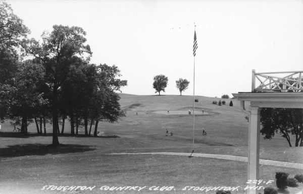 Text on front reads: "Stoughton Country Club, Stoughton, Wis." Golfers are playing on the course on a hill with trees and shrubs. The entrance to a building is in the right foreground and an American flag in the center foreground.