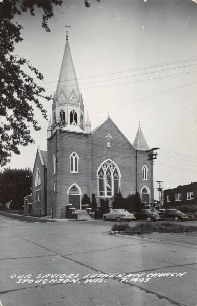 Text on front reads: "Our Saviors Lutheran Church, Stoughton, Wis." The Neogothic Revival style church was constructed of concrete block in 1904. Also known as the Old North Church, it was demolished in 1996. It has arched stained glass windows and doorways and an ornate belfry. Automobiles are parked in front.