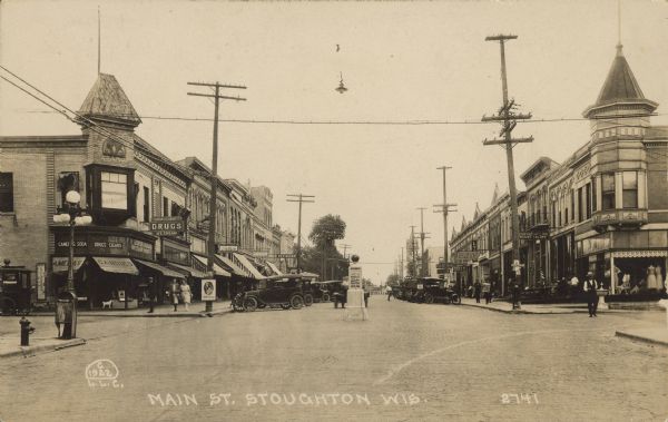 Text on front reads: "Main St. Stoughton, Wis." A busy brick paved street lined with sidewalks and businesses. Automobiles are parked at the curb and pedestrians are on the sidewalks. A dog is standing in the drugstore door on the left. Some of the signs read: "Cameras, Soda, Drugs, Cigars, Candies, Ice Cream, Restaurant, Anderson Shoes, Furniture, Cafe and Tailors." A marker in the middle of the intersection reads: "Division Street, Service, Garage, Agency, Nash Six and and Motor Cars" with an arrow pointing to the right.
