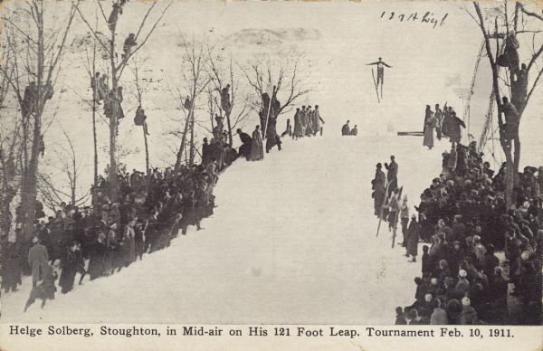 Text on front reads: "Helge Solberg, Stoughton, in Mid-air on his 121 Foot Leap. Tournament Feb. 10, 1911." Helge Solberg ski jumping at a tournament, the crowd of spectators watching from the sides, and with some people perched in the trees.