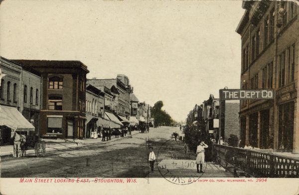 Text on front reads: "Main Street Looking East, Stoughton, Wis." A busy  street lined with sidewalks and businesses. Horse-drawn vehicles are pulled to the curb and pedestrians are on the sidewalks. On the right is a sign hanging over the sidewalk that reads: "The Dept Co", with an iron fence and wooden platform below it. Two children are standing on the platform.