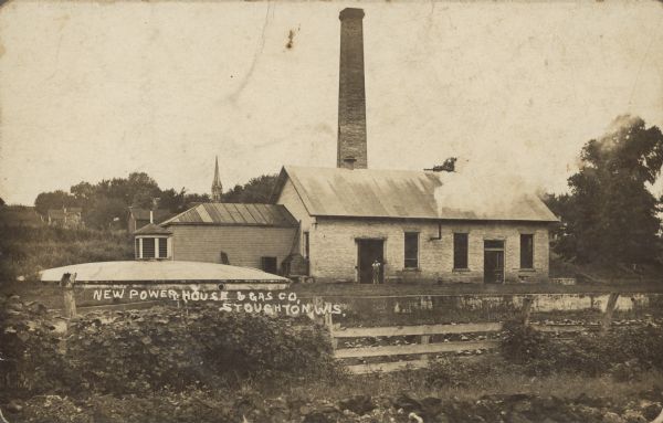 Text on the front reads: "New Power House & Gas Co, Stoughton, Wis." A brick building with a wooden addition, with a man standing in the doorway. Steam is billowing out of a pipe protruding through the wall. On the left is a semi-underground tank with a cupola, behind a concrete retaining wall. In the foreground is a fence with vegetation and rocks. In the background is a tall chimney, buildings, trees, and a church steeple.