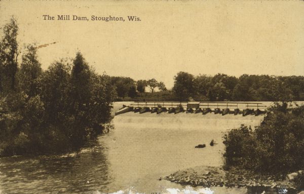 Text on front reads: "The Mill Dam, Stoughton, Wis." Elevated view towards the dam with trees and shrubs on the shoreline.