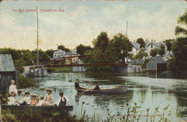 Text on front reads: "The Old Bridge, Stoughton, Wis." View along shoreline towards a boat on the Yahara River with two boys in it, and another boat at the shore with eight children in it. In the background a man is standing on the approach to the metal bridge spanning the river. A boathouse is on the left and a neighborhood is on the right.
