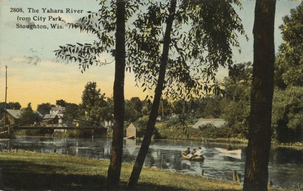 Text on front reads: "The Yahara River from City Park, Stoughton, Wis." View from shoreline towards a river flowing through a town park. There are two boats on the river, one with a single passenger and the other with three. The shoreline is lined with trees, foliage and a boathouse. In the background are a metal bridge, and beyond are dwellings of a neighborhood among trees.