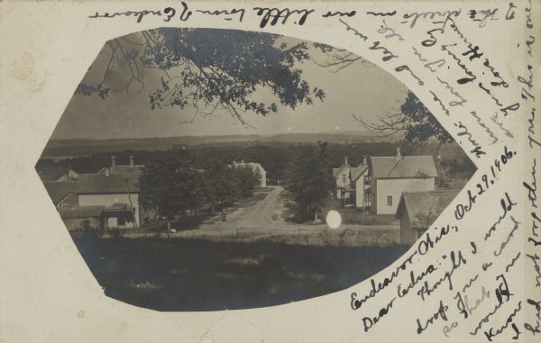 View from a hillside towards a woman walking on an unpaved neighborhood street in a small town, with dwellings and trees on both sides. Along the horizon are tree-covered hills. The image has an interestingly shaped mask on it, and the writer arranged her message to match.