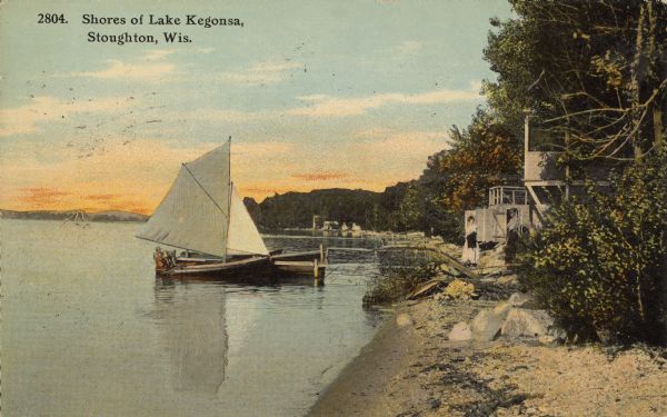 Text on front reads: "Shores of Lake Kegonsa, Stoughton, Wis." A man and woman are standing on the shoreline while three children are sitting in a sailboat tied at the dock. Along the shoreline are boathouses, cottages, trees and docks. A swimming structure is further out from shore in the left background.
