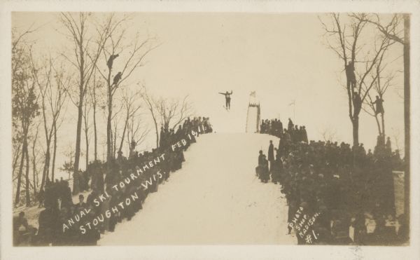 Text on front reads: "Anual [sic] Ski Tournament, Feb 10, 11. Stoughton, Wis." Ski jumping at a tournament with the crowd of spectators watching from the sides and perched in the trees. Handwriting on the reverse: "The longest standing jump was 135 feet. It looks best in a picture because you do not have to freeze to see it."