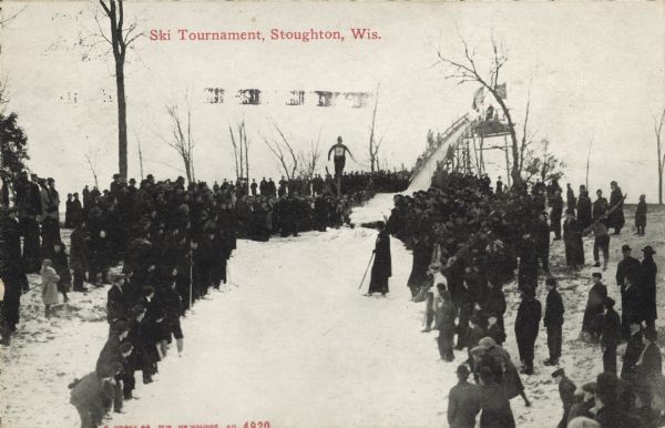 Text on front reads: "Ski Tournament, Stoughton, Wis." A ski jumper in mid-air at a tournament, with the crowd of spectators watching from the sides.