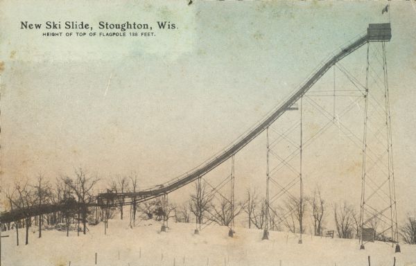 Text on front reads: "New Ski Slide, Stoughton, Wis. Height of Top of Flagpole 138 Feet." The side view of a jumping ramp, also called an in-run, at a ski hill. Snow covers the ground with trees on the horizon.