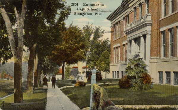 Text on front reads: "Entrance to High School, Stoughton, Wis." Four teenage girls pose on the walk to the entrance of the High School, and more students are on the sidewalk below. Plants landscape the lawn, mature trees line the street and a fence and rock are in the foreground.