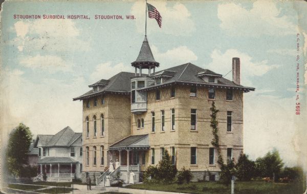 Text on front reads: "Stoughton Surgical Hospital, Stoughton, Wis." Two women are standing on the porch and a man and two more women are standing on the sidewalk below. The building is brick with 3 stories and a basement level. A tower on the roof is flying an American Flag. Homes are on the left.