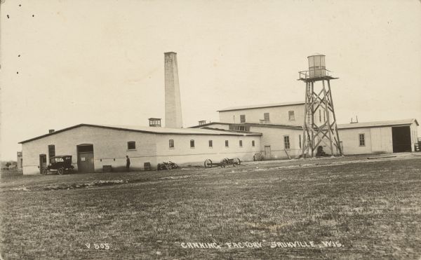Text on front reads: "Canning Factory, Saukville, Wis." Brick factory buildings with a tall chimney and a water tower. A man and an automobile can be seen at the end of the building on the left, a wagon chassis in the center.