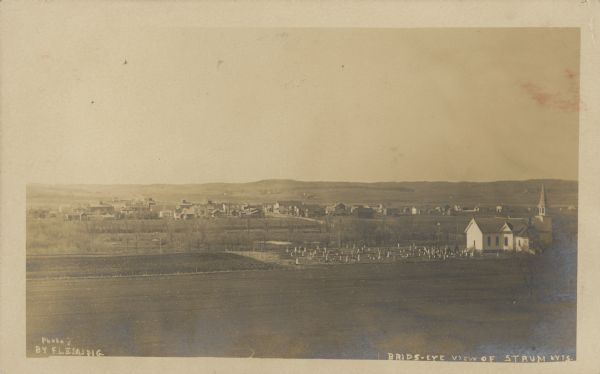 Text on front reads: "Bird's Eye View of Strum, Wis." Elevated view of a small town with hills on the horizon. A church and cemetery can be seen on the right with tilled fields in the foreground.