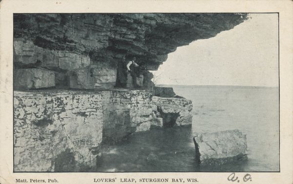 Text on front reads: "Lovers' Leap, Sturgeon Bay, Wis." Two men are posing while sitting on a block of stone under an overhang on Cabot's Point. The shoreline is rocky and jagged.