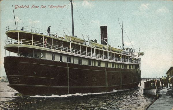 Text on front reads: "Goodrich Line Str. 'Carolina'." "Carolina of Duluth" is painted on the prow of the steamer. Passengers are lined up along the railings of the decks with lifeboats on the top deck. A pier, a smaller boat and people are on the right. A bridge and shoreline are on the horizon.