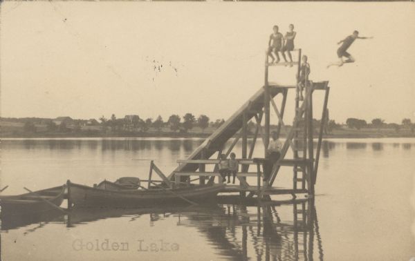 Text on front reads: "Golden Lake." A man and six children posing on a dock with a water slide and diving platform. One of the boys is leaping into the water. Three rowboats are tethered to the dock. Buildings and trees are on the far shore. Golden Lake is located on the shared boundary between Jefferson and Waukesha counties.