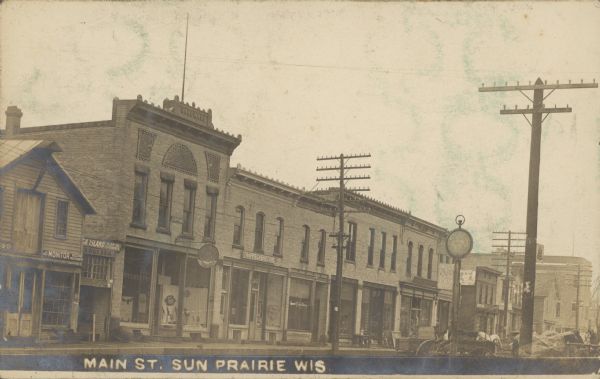 Text on front reads: "Main St. Sun Prairie Wis." View of storefronts line the left side of the street, with horse-drawn wagons at the curb. A man in an apron is standing on the sidewalk. Some of the signs read: "Monitor Seeding Machinery", "Rock Island Plow Co. Implements", "Hardware", "Favorite Stove and Ranges", "Trapps Cash Store", "Bank of Sun Prairie", "Engel Brothers", "Monarch Paint" and "Parker Fountain Pen."