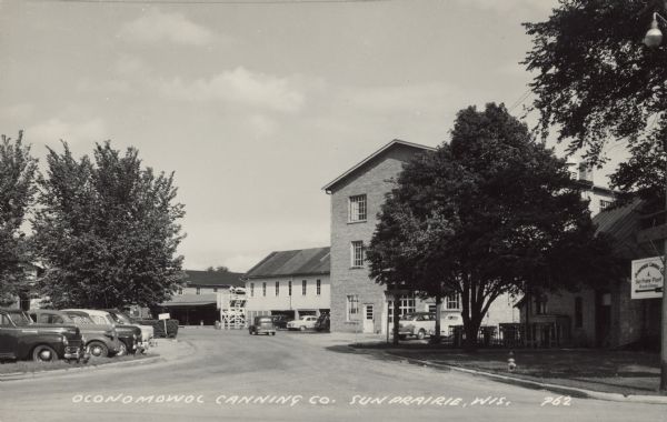 Text on front reads: "Oconomowoc Canning Co. Sun Prairie, Wis." A group of buildings at a canning factory. A sign on the right reads: "Oconomowoc Canning Company, Sun Prairie Plant, Private Entrance." The two buildings on the right are built of brick. There are trees and parked automobiles.