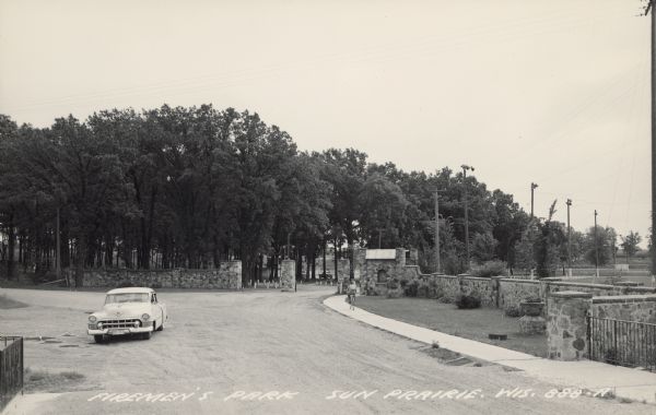 Text on front reads: "Fireman's Park, Sun Prairie, Wis." The entrance to a park with stone walls, sidewalks and trees. A Cadillac is parked in the street and a man on a bicycle rides into the park.
