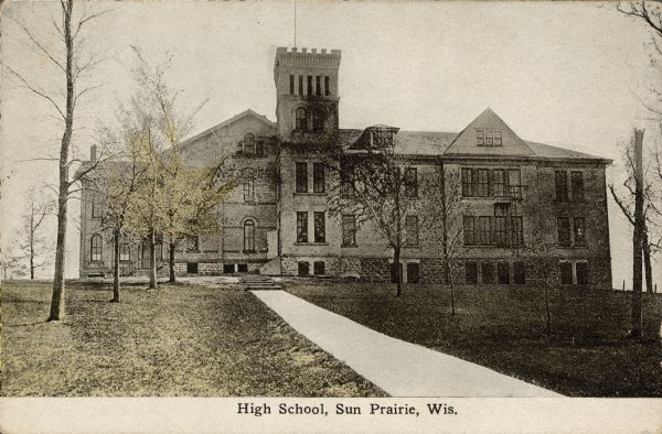 Text on front reads: "High School, Sun Prairie, Wis." A brick and stone building with many windows and a square tower. Trees and a sidewalk lead to the entrance.
