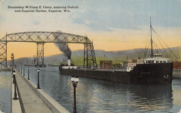 Text on front reads: "Steamship William E. Corey, entering Duluth and Superior Harbor, Wis." A steamship entering the harbor under the Aerial Lift Bridge, completed in 1905. It was remodeled and reopened in 1930.  A row of lampposts light the pier, with a beacon or lighthouse in the distance. Buildings are on the right with hills in the background.