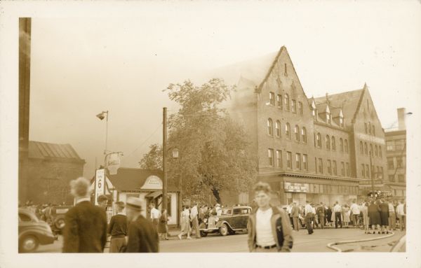 Crowds stand in the street watching as a fire burns in the four story, brick Hotel Superior. Automobiles are parked at the curb and more people are walking towards the fire. Nine fire companies attempted to stop the blaze. Signs read: "Hotel Superior", "Sylvia's Children Shop", "Goodyear Shoe Works" and "Shell."
