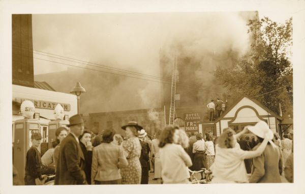 A crowd of people are standing in the street watching as a fire burns in the four story, brick Hotel Superior and a dozen stores and offices. Nine fire companies attempted to stop the blaze. There is a Shell Station on the left and two men are standing on the roof of the Goodyear Shoe Works. A sign on the hotel reads: "Hotel Superior, Free Parking."