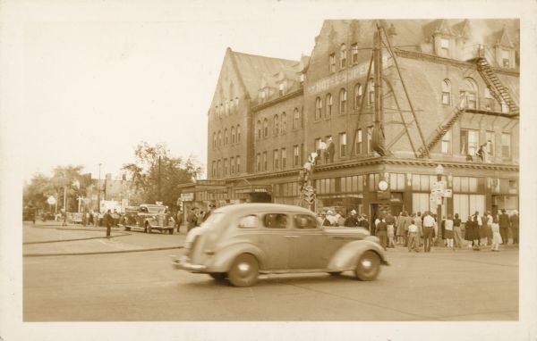 Crowds standing in the street watching as a fire burns in the four story, brick Hotel Superior. People are being rescued with a ladder and others have escaped out a window and are standing on the ledge. An automobile is driving through the intersection and firetrucks are parked on the left. Nine fire companies attempted to stop the blaze.