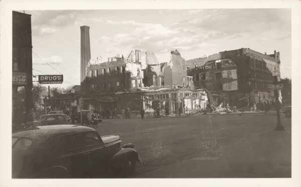 A crowd of people view the aftermath of a fire that destroyed the four story brick Hotel Superior and a dozen stores and offices. Nine fire companies attempted to stop the blaze. A sign on the left reads: "Drugs, John S. Hadley." Automobiles are in the street and parked at the curb.