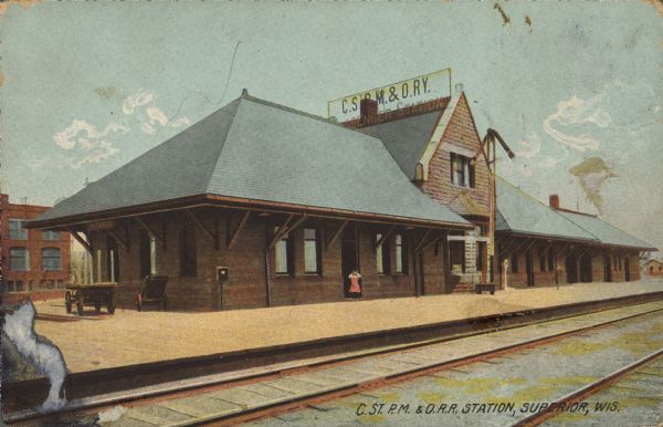 Text on front reads: "C.ST.P.M. & O.R.R. Station, Superior, Wis." A girl stands in the doorway of the Chicago, St. Paul, Minneapolis and Omaha Railway train station. In the foreground is the loading platform and railroad tracks. On the left are two baggage carts, and buildings in the background. The sign reads: "C.ST.P.M. & O.R.R. Messenger Station."