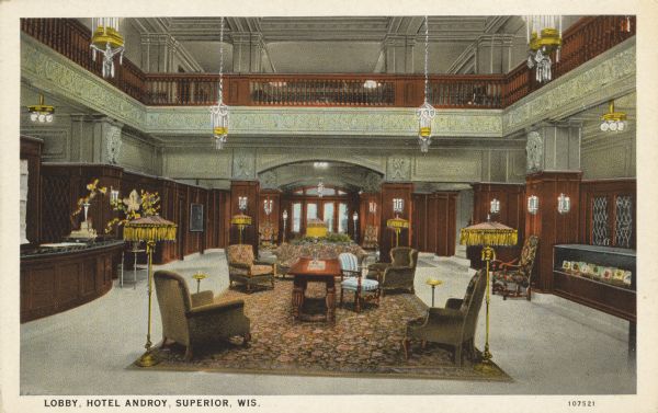 Text on front reads: "Lobby, Hotel Androy, Superior, Wis." An ornately decorated space with wooden paneling, hanging lights and sconces on the walls. Fringed lamps and upholstered seating form a seating area in the center. The reception area is on the left.