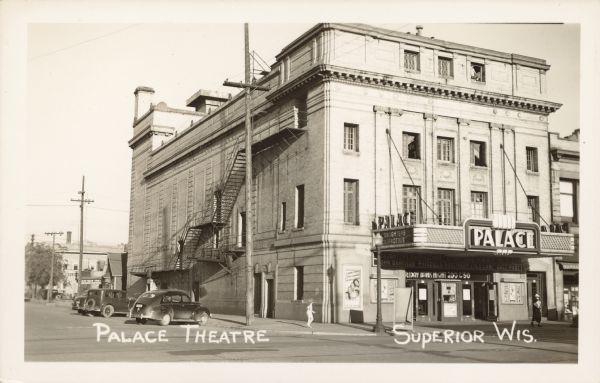 Text on front reads: "Palace Theatre, Superior, Wis." A brick theatre with many windows and ornate decorative elements on the exterior. A child is on the sidewalk in the center and a woman on the right. A man is perched in a 4th story window. Automobiles are parked at the curb on the left. Text on the posters, front and marquis read: "Daughters Courageous, Everything New but the Cast, John Garfield, Priscilla Rosemary, Lola Lane, Gale Page", "Friday Bank Night 200&50." The theatre was opened in 1917, remodeled in 1953, closed in 1982 and torn down in 2006.
