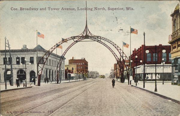Text on front reads: "Cor. Broadway and Tower Avenue, Looking North, Superior, Wis." The Grand Army of the Republic Arch spans the corner of Broadway and Tower Avenue. Built in 1900 to honor Civil War veterans, it contained 220 light bulbs at one time and was taken down in 1921 due to age. Two sets of tracks run under the arch with streetcars in the distance. Businesses and lampposts line the streets and pedestrians are walking in the street and on the sidewalks. Two signs read: "Coal" and "Closing Out Sale."