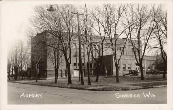 Text on front reads: "Armory, Superior, Wis." Corner view of a four-story brick building surrounded by trees. Displayed on the lawn are two cannons and a monument.