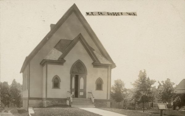 Text on front reads: "M.E. Ch. Sussex, Wis." A Gothic Revival church built in 1894. A sign is near the sidewalk in the right foreground, and a dwelling and fence in the right background. The church is still in use today.