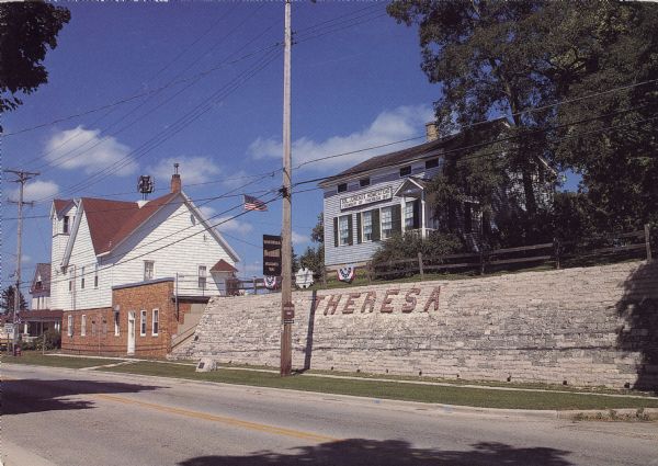 Text on reverse reads: "Theresa, Wisconsin 53091. The Old Stone Wall, Old Village Hall, and Solomon Juneau Homestead are landmarks in the picturesque, little village of Theresa." The clapboard Solomon Juneau Homestead was built in 1847 in the Greek Revival Style. In June, 1932, the house was moved to this location from its original site, higher up on the hill. The Village Hall was built in 1900. The name "THERESA" is built into the wall. Signs read: "Sol. Juneau's Homestead, Founder of Theresa 1847" and "Theresa Welcomes You."