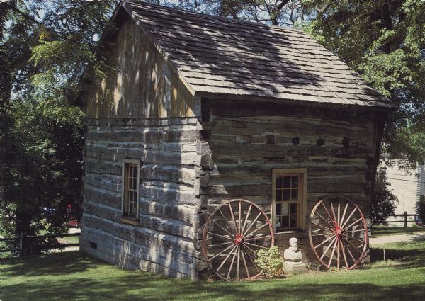 Text on reverse reads: "Theresa, Wisconsin 53091. The Reklau log house is displayed and maintained by Theresa Historical Society. The house was built about 1850 and was located 1 1/2 miles south of Theresa. In 1990, it was moved to its present site in the Theresa Historical Park." A log cabin moved from its original site to a historical park, surrounded by trees. Two wagon wheels and the bust of a man are on display on the side.