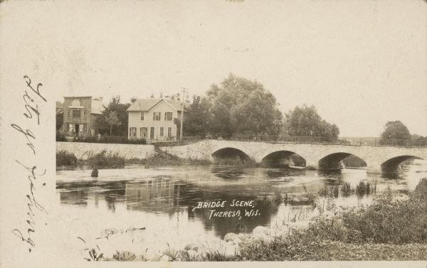 Text on front reads: "Bridge Scene, Theresa, Wis." The stone arch bridge over the East Branch Rock River was built in 1874, replacing the original bridge built in 1951. It was replaced in 1971. A dwelling and a business can be seen on the left.