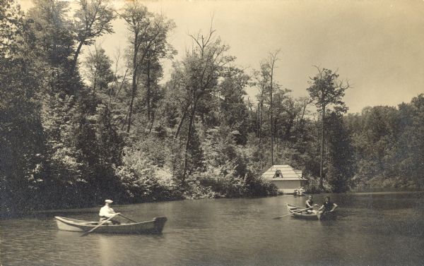 Handwriting on reverse reads: "Bornstein's boat house (formerly Blommer) and our 3 mile lake created by the dam." Three men are in two rowboats on a lake. Another boat is located near the boathouse on the right. The lake is surrounded by trees.