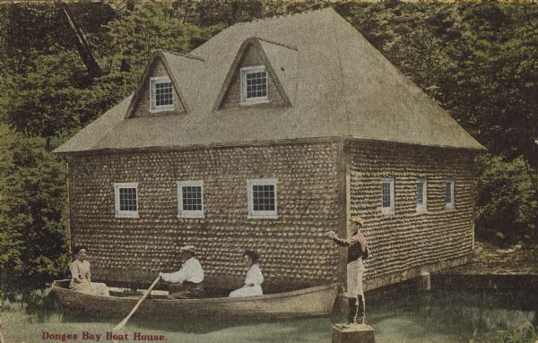 Text on front reads: "Donges Bay Boat House." A man rowing a boat with two women passengers, in front of a boathouse built of rocks. A lawn jockey stands on a plinth in the water. The background is filled with trees.