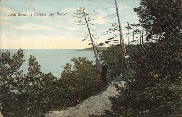 Text on front reads: "John Schuch's Donges Bay Resort." A man stands on a trail, gazing over shrubs at a bay on Lake Michigan. The shoreline is surrounded by trees.