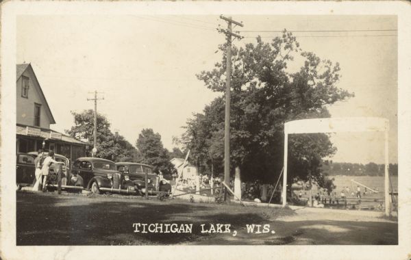 Text on front reads: "Tichigan Lake, Wis." A couple stands next to parked cars in front of the grocery store at Wichmans Resort. On the right is a crowd of people near the cottages on the lake, on the docks  and in the water. Trees and docks line the shore. On the far right is a large overhead sign. Signs read: "Wichmans Resort", "Grocery" and "Schlitz on Tap."