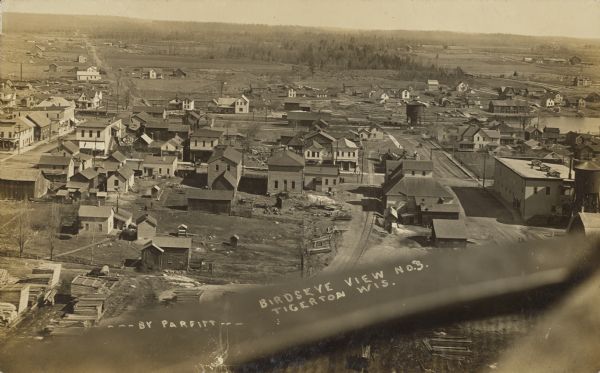 Text on front reads: "Birdseye View, Tigerton, Wis." View of the town with railroad tracks and unpaved streets. Many streets have sidewalks or boardwalks, with a few pedestrians. Wooded hills are along the horizon. A railing or other object is in the foreground.