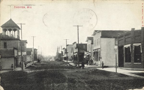 Text on front reads: "Tigerton, Wis." Pedestrians are on the sidewalks, boardwalks and in the unpaved street of the town. Storefronts and businesses line the street and horse-drawn vehicles are at the curb. The building on the left corner has a bell tower.