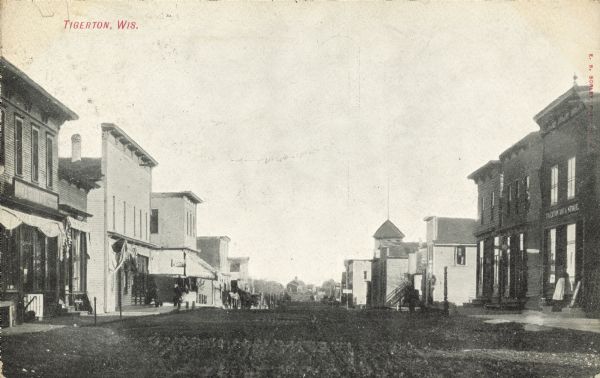 Text on front reads: "Tigerton, Wis." Storefronts and businesses line the unpaved street. A woman is standing on the stoop in front of a store on the right. 