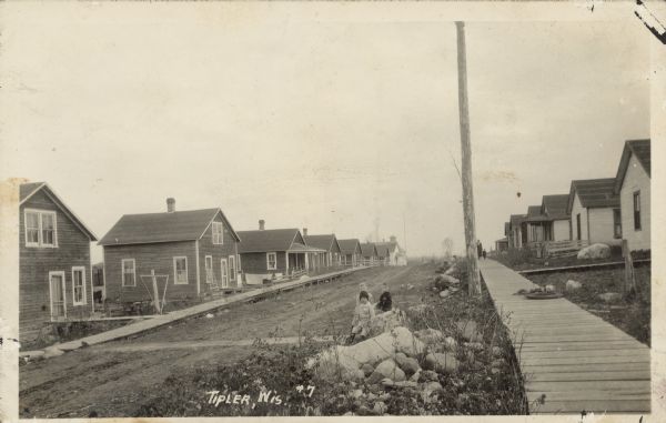 Text on front reads: "Tipler, Wis." Many clapboard homes line an unpaved street with boardwalks in a residential neighborhood. Children are sitting on a rock pile in the foreground. An woman and child are walking on the boardwalk on the right.