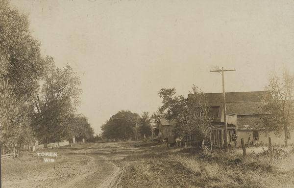 Text on front reads: "Token, Wis." Unpaved street with homes, barns, fences and trees. A horse and buggy are on the right.